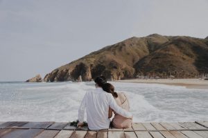 Pareja sentada en un muelle frente al mar y las montañas, abrazados. Amor y empatía para desarrollar la conciencia del mundo.