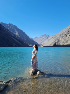 Mujer en equilibrio sobre las rocas, contemplando el lago sereno entre majestuosas montañas. En este paisaje tranquilo, ella busca el despertar de la conciencia espiritual, conectándose con la paz interior y la sabiduría que emana de la naturaleza.