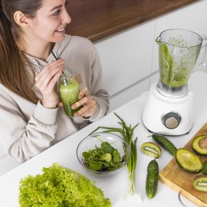 Mujer sonriente preparando un jugo verde detox con kiwi, aguacate, pepino y lechuga. La dieta para desintoxicar el cuerpo incluye alimentos frescos y naturales para mejorar la salud y el bienestar.