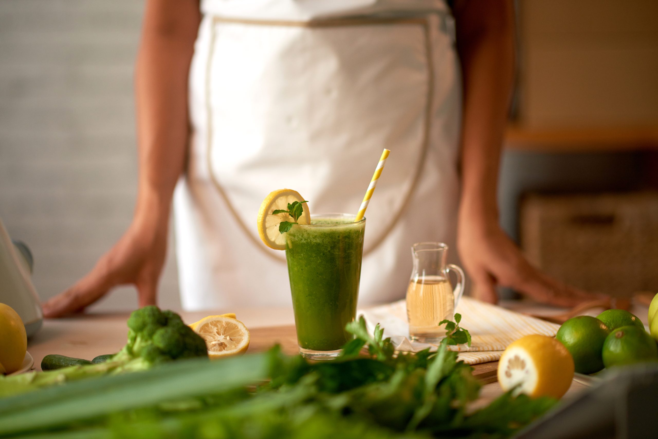 Un vaso de jugo verde detox con rodaja de limón, rodeado de brócoli, limón y apio sobre una mesa de madera. La dieta para desintoxicar el cuerpo incluye alimentos frescos y naturales para mejorar la salud y el bienestar.
