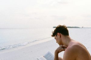 Joven leyendo un libro en la playa, frente al mar. Exploración y aprendizaje para aumentar la conciencia del mundo.