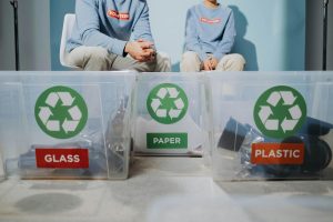 Contenedores de reciclaje con etiquetas de vidrio, papel y plástico, con dos personas clasificando materiales al fondo.