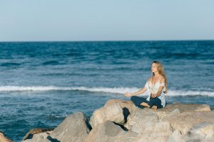 Mujer meditando en armonía sobre rocas y de fondo el imponente mar. 