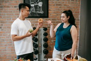 Joven sosteniendo una dona y una manzana, con una chica señalando la dona. Pesas de fondo. Concepto de elección de alimentos saludables frente a indulgencias. Nutrición y deporte para una vida equilibrada y activa.