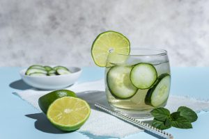 Un vaso de agua infusionada con rodajas de pepino, limón y hojas de menta, colocados en una mesa de cocina con luz natural. 