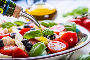Variedad de alimentos frescos de la dieta mediterránea siendo rociados con aceite de oliva, ejemplificando qué es la dieta mediterránea.