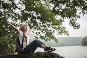 mujer joven sonriendo y disfrutando de la naturaleza, sentada sobre una roca con una mochila a su lado. La imagen transmite la importancia de desconectar de la tecnología y reconectar con la naturaleza para mejorar el bienestar digital.
