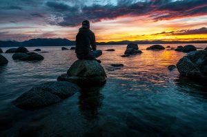 Persona sentada en una roca junto al agua, reflexionando sobre el autoconocimiento y el crecimiento personal en un entorno natural de montañas y ríos.