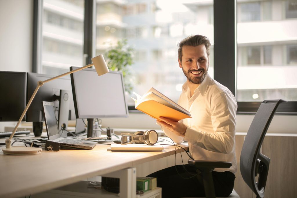 Emprendedor concentrado frente a su ordenador, buscando inspiración en un libro mientras sonríe con confianza. El desarrollo personal para emprendedores impulsa su camino hacia el éxito.