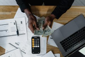 Manos contando dólares, rodeadas de una computadora, calculadora y anotador. Esta imagen destaca la importancia del desarrollo personal en el éxito financiero del emprendedor. ¡Descubre cómo el crecimiento personal impulsa tus metas financieras!
