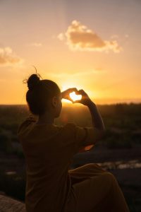 Mujer mostrando un corazón con el sol brillando en el cielo, simbolizando amor y conexión con la naturaleza.